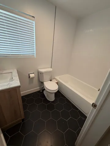 Bathroom featuring dark hexagon floor tiles, a white bathtub, a toilet, a vanity with a white countertop and integrated sink, and a window with blinds