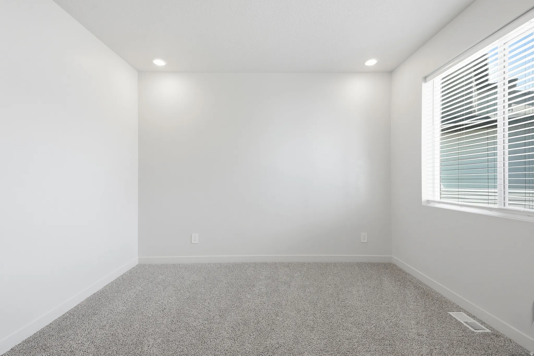Bright room featuring light gray carpeting, white walls, recessed ceiling lighting, and a window with horizontal blinds