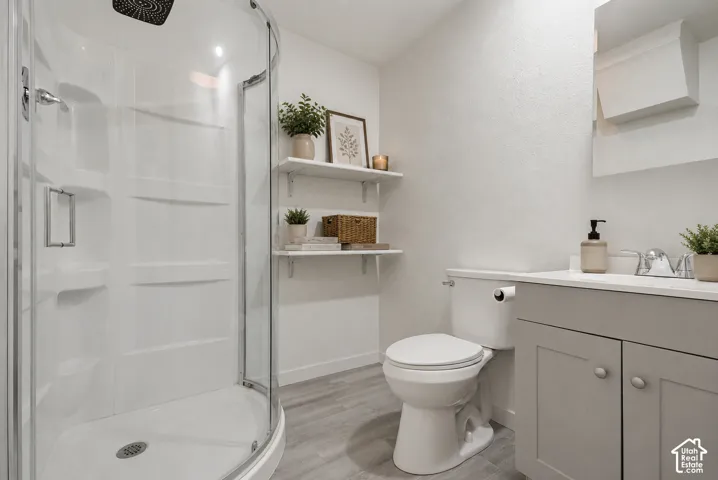 Three-piece bathroom featuring a curved glass shower enclosure, a single vanity with a light gray cabinet, a white countertop, and wood-finish flooring