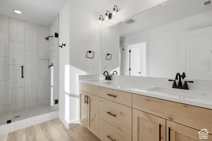Dual vanity with undermount sinks and light wood-finish cabinetry, quartz-finish countertop, matte black fixtures, full-width mirror, and a glass-enclosed shower with tile surround