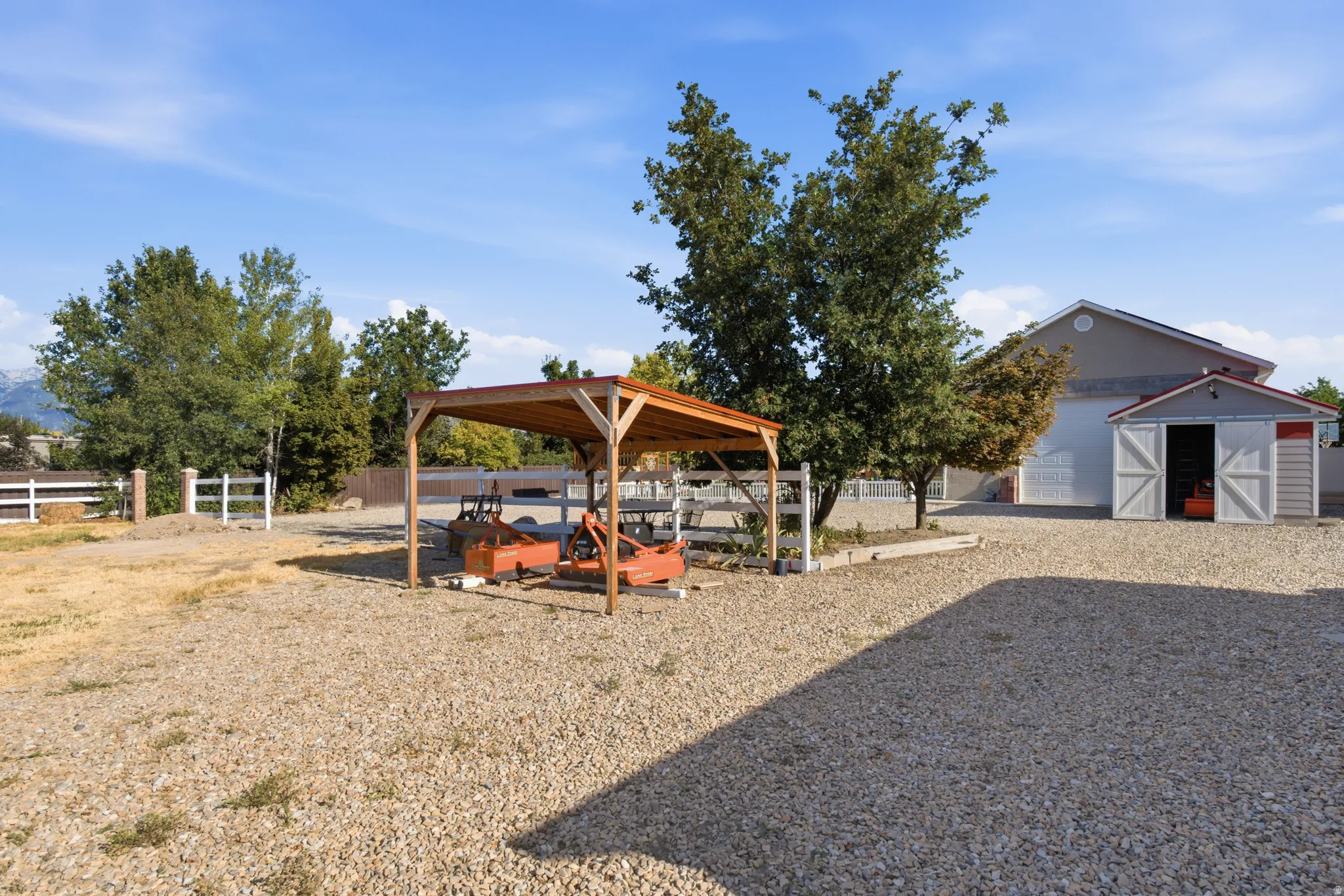 Gravel driveway with a freestanding wood-frame carport, a white picket fence, a small storage shed, and mature trees