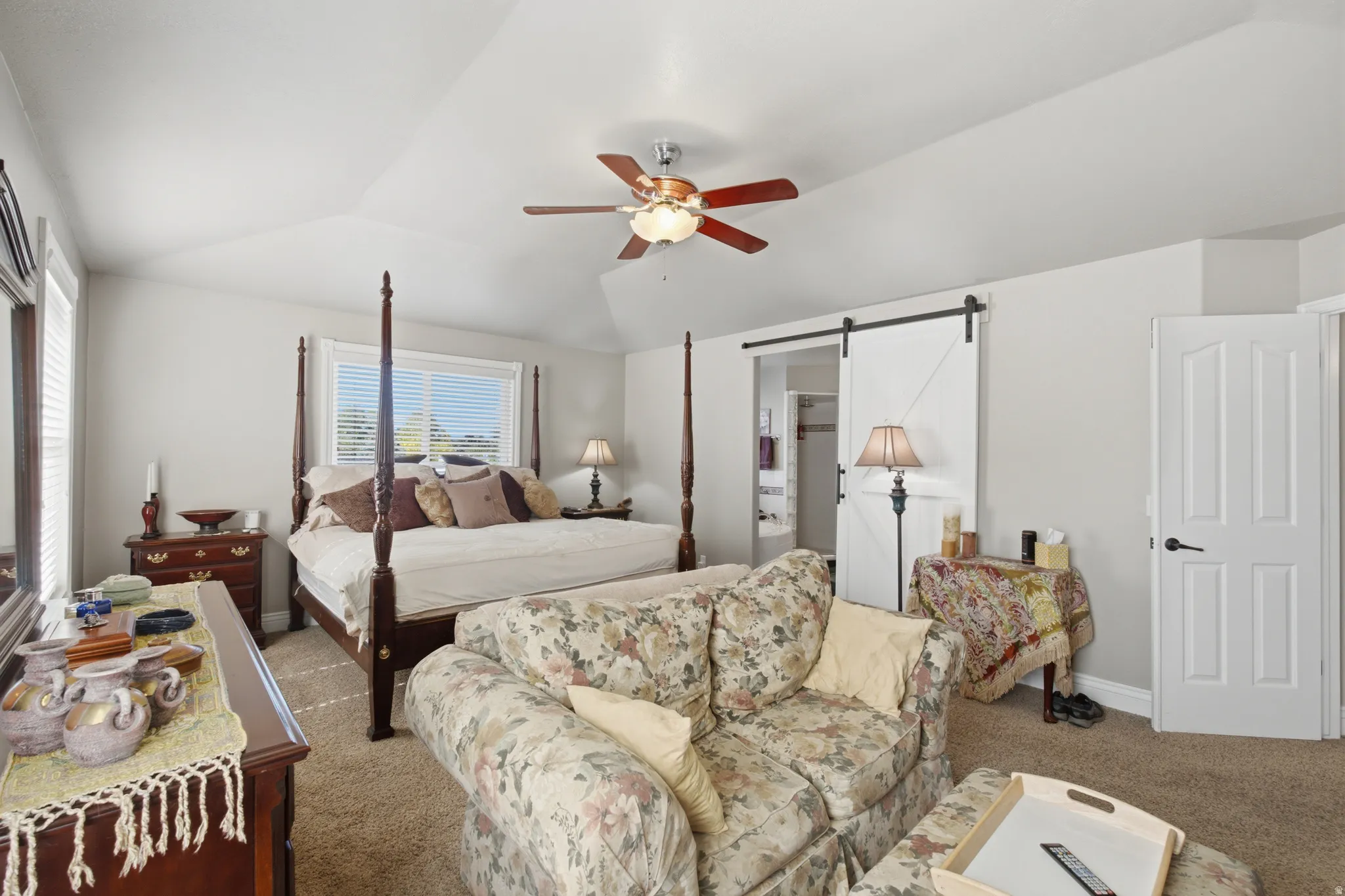 Spacious bedroom featuring vaulted ceilings, a window with horizontal blinds, neutral carpet flooring, and a white barn door
