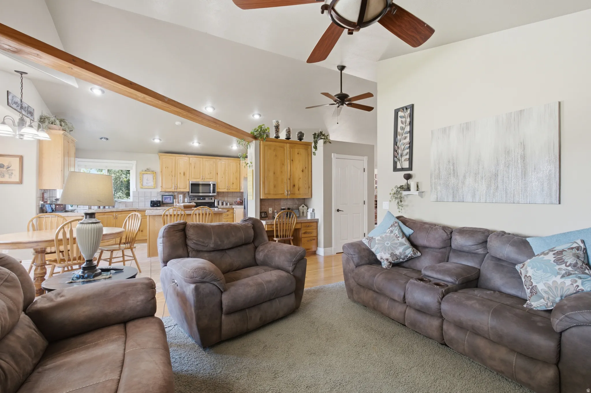 Open-concept living area featuring vaulted ceilings, a prominent wood beam, ceiling fans, recessed lighting, and a kitchen with wood cabinetry and a dining area