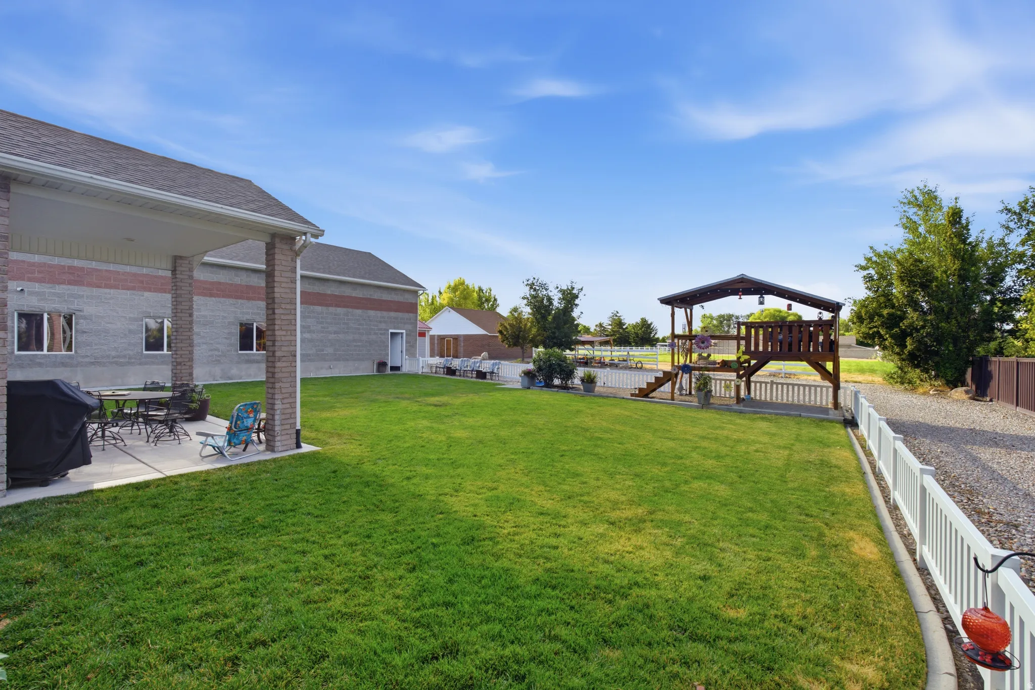 Expansive green lawn featuring a covered patio, a multi-level outdoor play structure, a gravel driveway, and a white picket fence