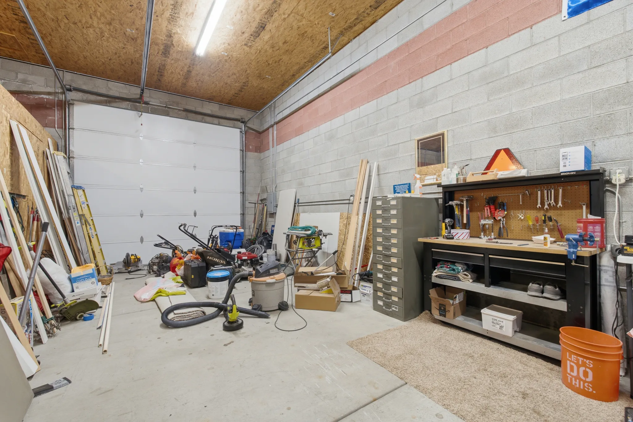 Spacious utility area featuring a large overhead door, concrete flooring, concrete block walls, wood-finish ceiling, and fluorescent lighting