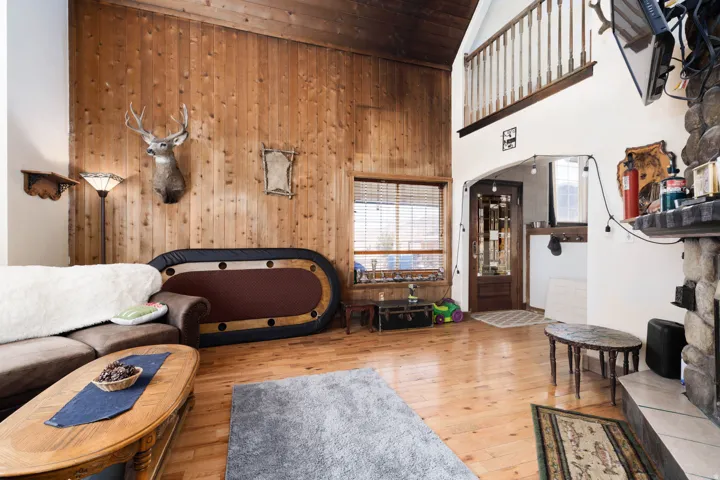 Vaulted ceiling great room featuring natural wood paneling, wood-finish flooring, a stone fireplace, an arched entryway, and a wood staircase with balusters