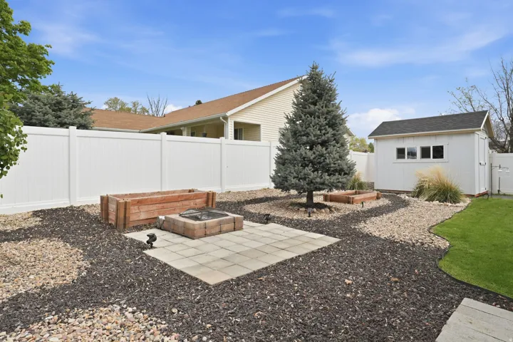 Expansive backyard featuring a white vinyl privacy fence, a detached storage shed with multiple windows, a mature evergreen tree, and a paved patio area with a built-in fire pit