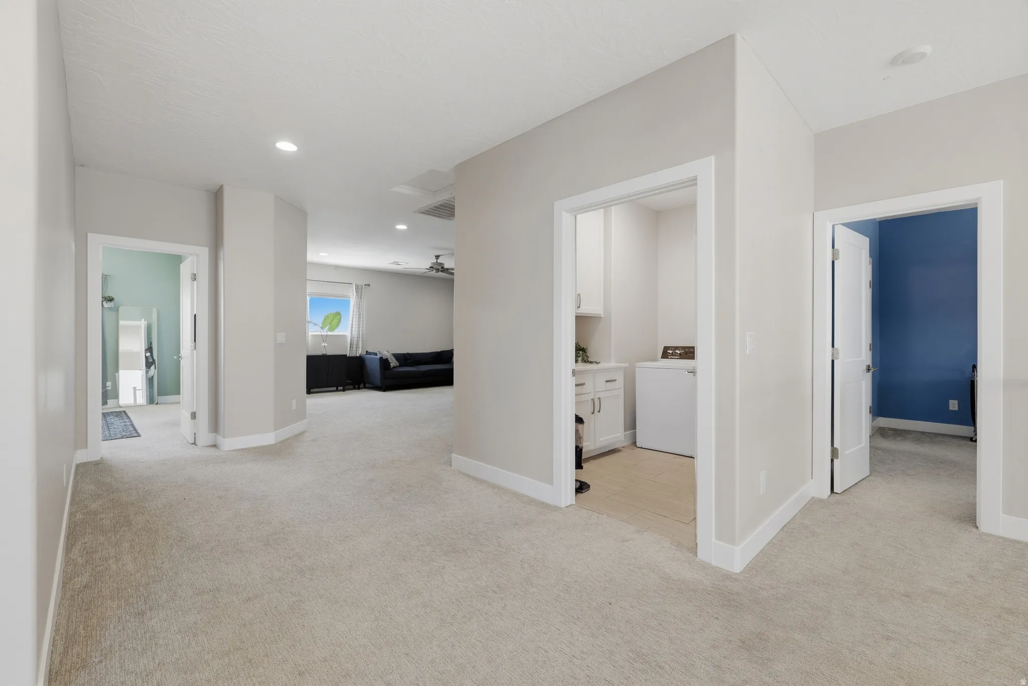 Spacious carpeted hallway with recessed lighting and white baseboards, offering access to a laundry area with white cabinetry, a room with a blue accent wall, and another room with light blue walls