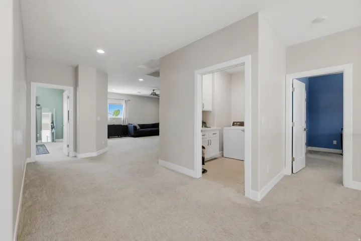 Spacious carpeted hallway with recessed lighting and white baseboards, offering access to a laundry area with white cabinetry, a room with a blue accent wall, and another room with light blue walls