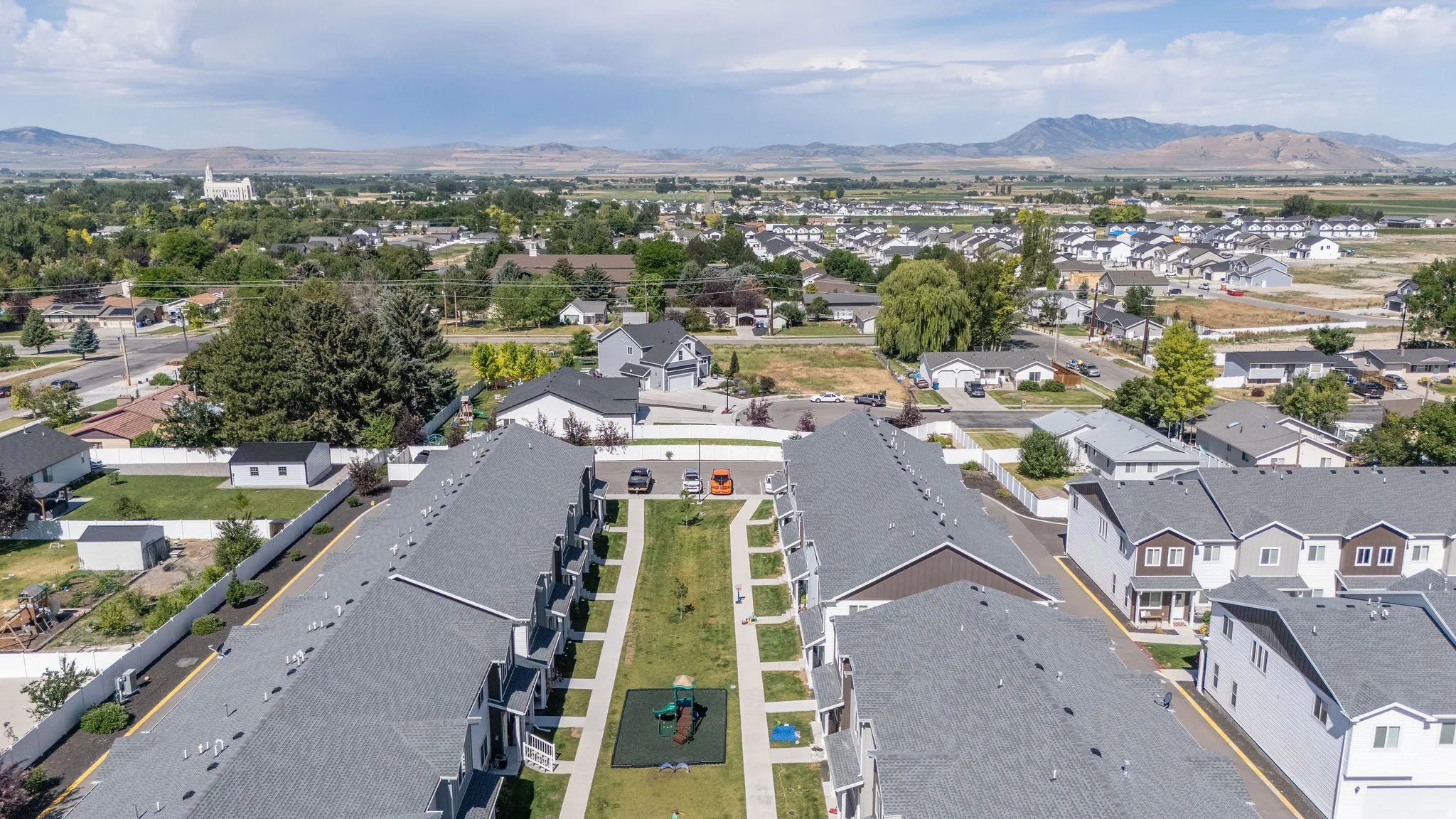 Aerial perspective showcasing a community of townhomes with gray shingle roofs and private patios, featuring a central green space with a playground