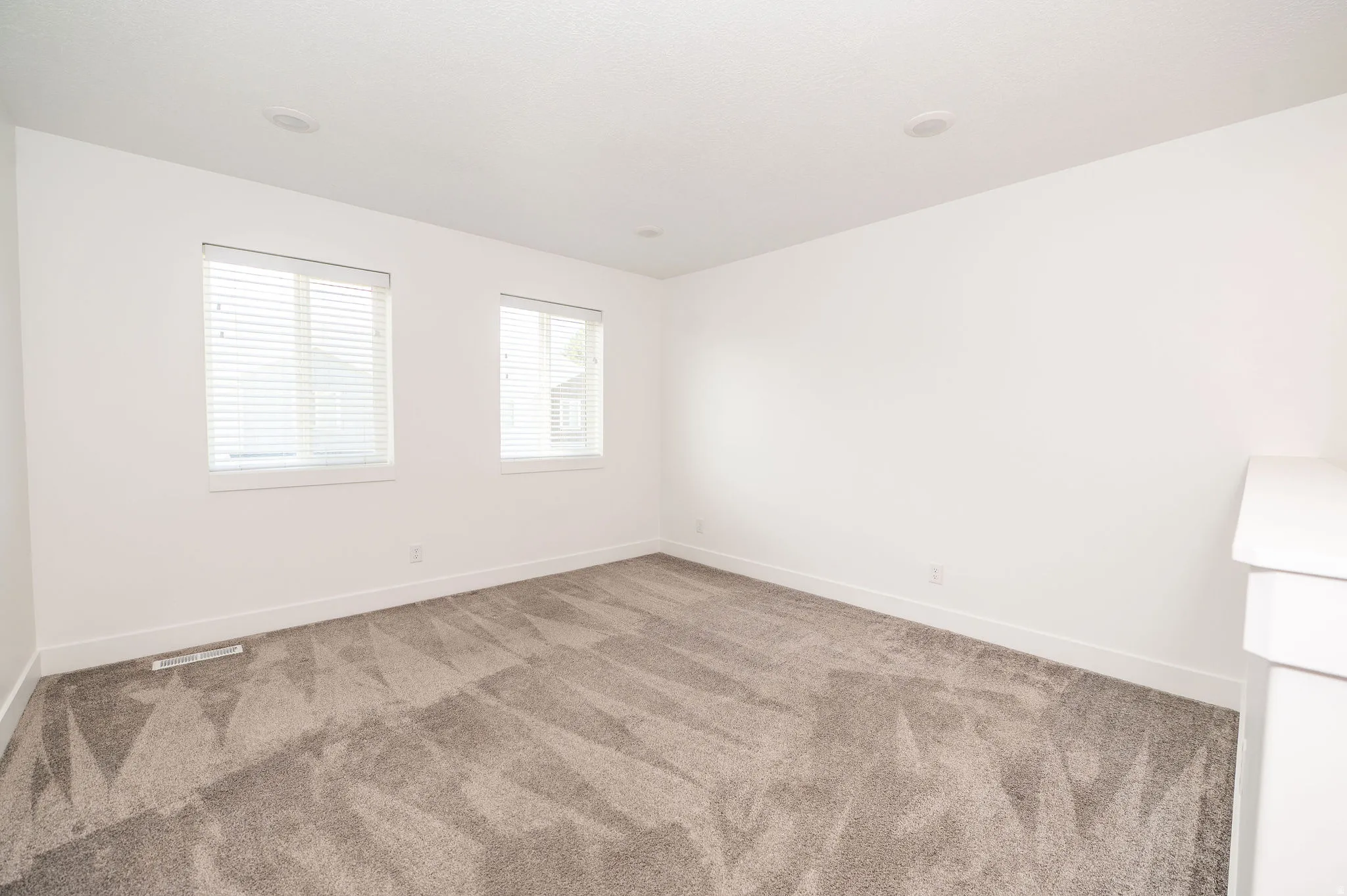 Spacious room featuring neutral carpeting, two windows with blinds, recessed lighting, and crisp white walls