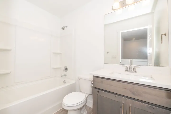 Full bathroom featuring a white tub and shower combination with integrated shelving, a sleek vanity with a light-toned countertop and an undermount sink, and wood-finish flooring