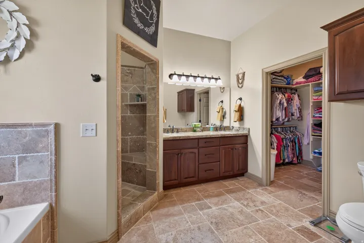 Expansive bathroom featuring large-format tile flooring, a walk-in shower with tiled walls and a built-in bench, a double vanity with a stone countertop and wood-finish cabinetry, and a spacious walk-in closet with built-in shelving and hanging rods
