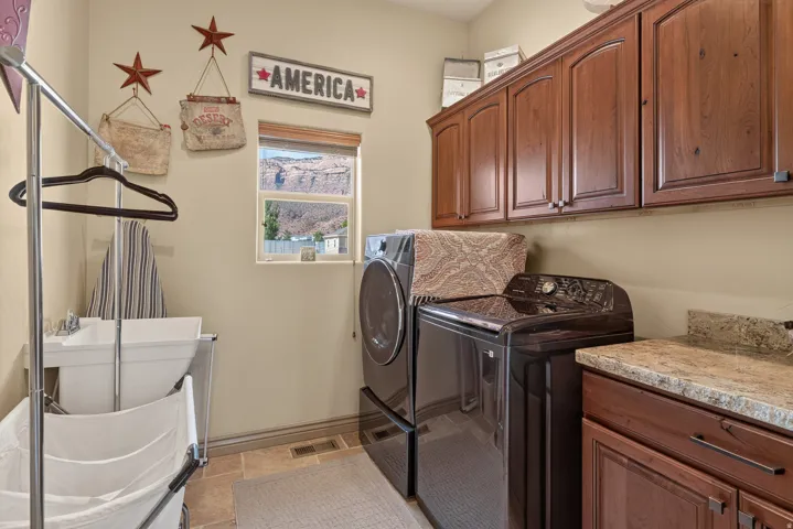 Utility room featuring upper and lower wood cabinetry, a window, a utility sink, and stone-finish countertops