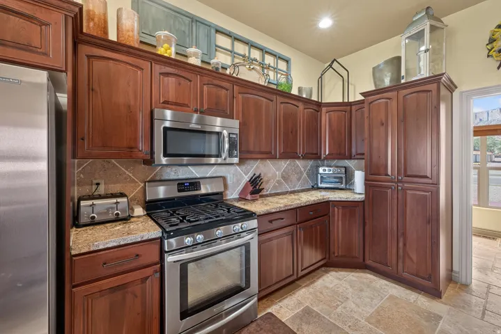 Kitchen featuring rich wood cabinetry, stainless steel appliances, patterned tile backsplash, natural stone countertops, and tile flooring