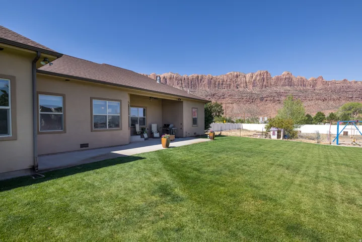 Expansive green lawn with a concrete patio under a stucco exterior, featuring multiple windows and a mountain backdrop