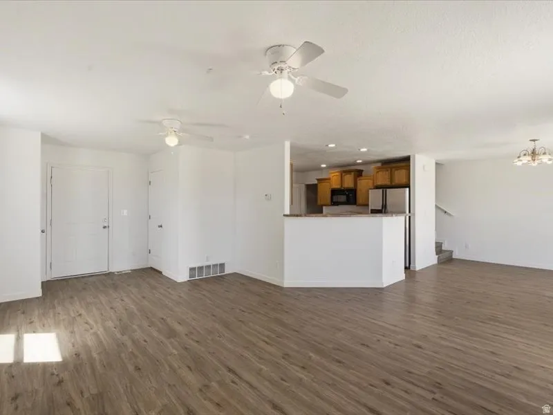 Open-concept living area featuring wood-finish flooring, white walls, two ceiling fans, and a kitchen visible with wood cabinetry, stainless steel appliances, and a light-colored countertop