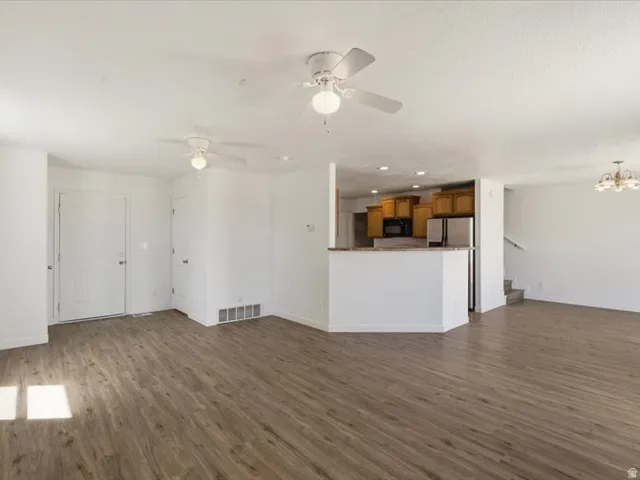 Open-concept living area featuring wood-finish flooring, white walls, two ceiling fans, and a kitchen visible with wood cabinetry, stainless steel appliances, and a light-colored countertop