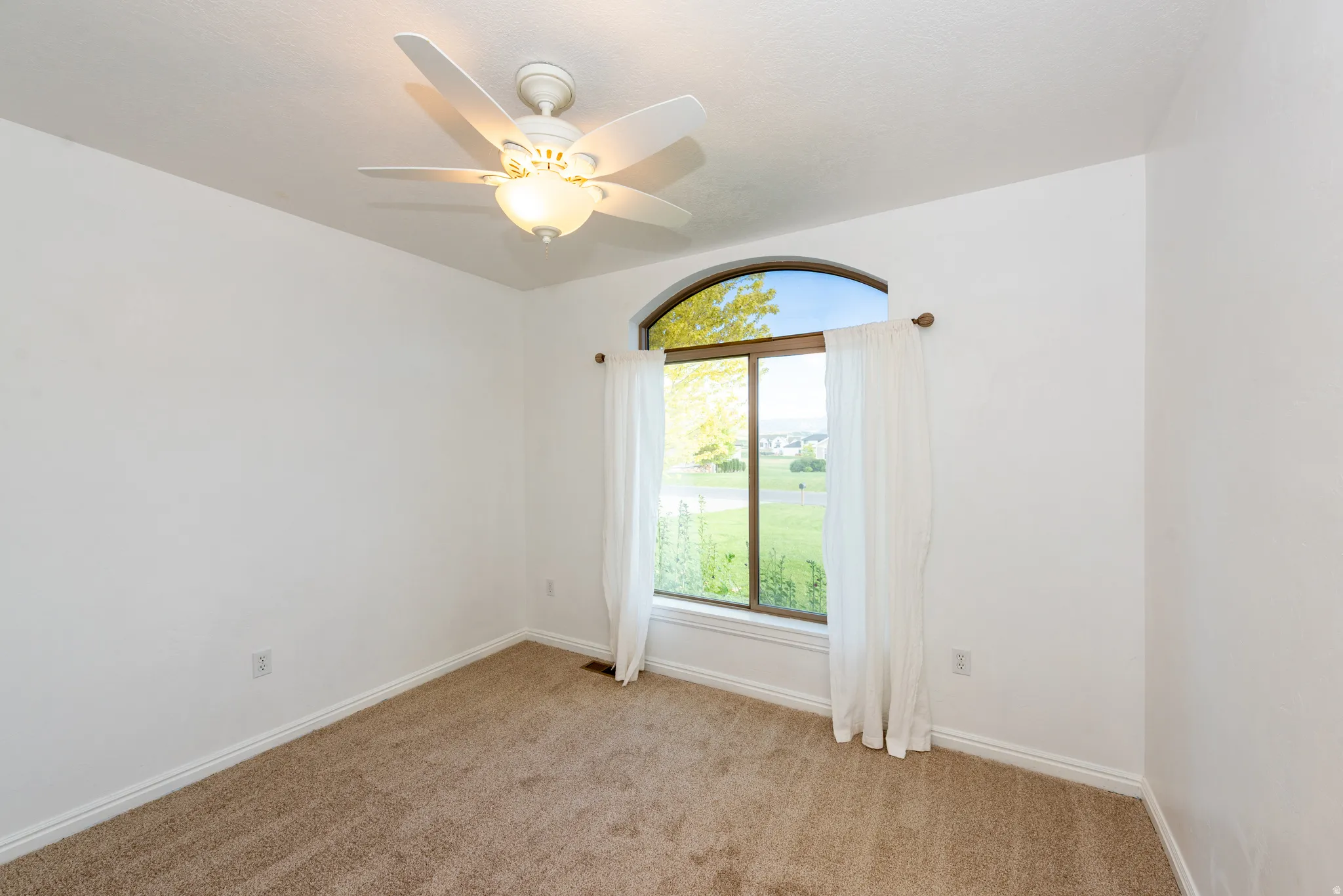 Room featuring a large arched window with a green outdoor landscape, neutral-toned carpeting, white baseboards, and a ceiling fan with integrated lighting