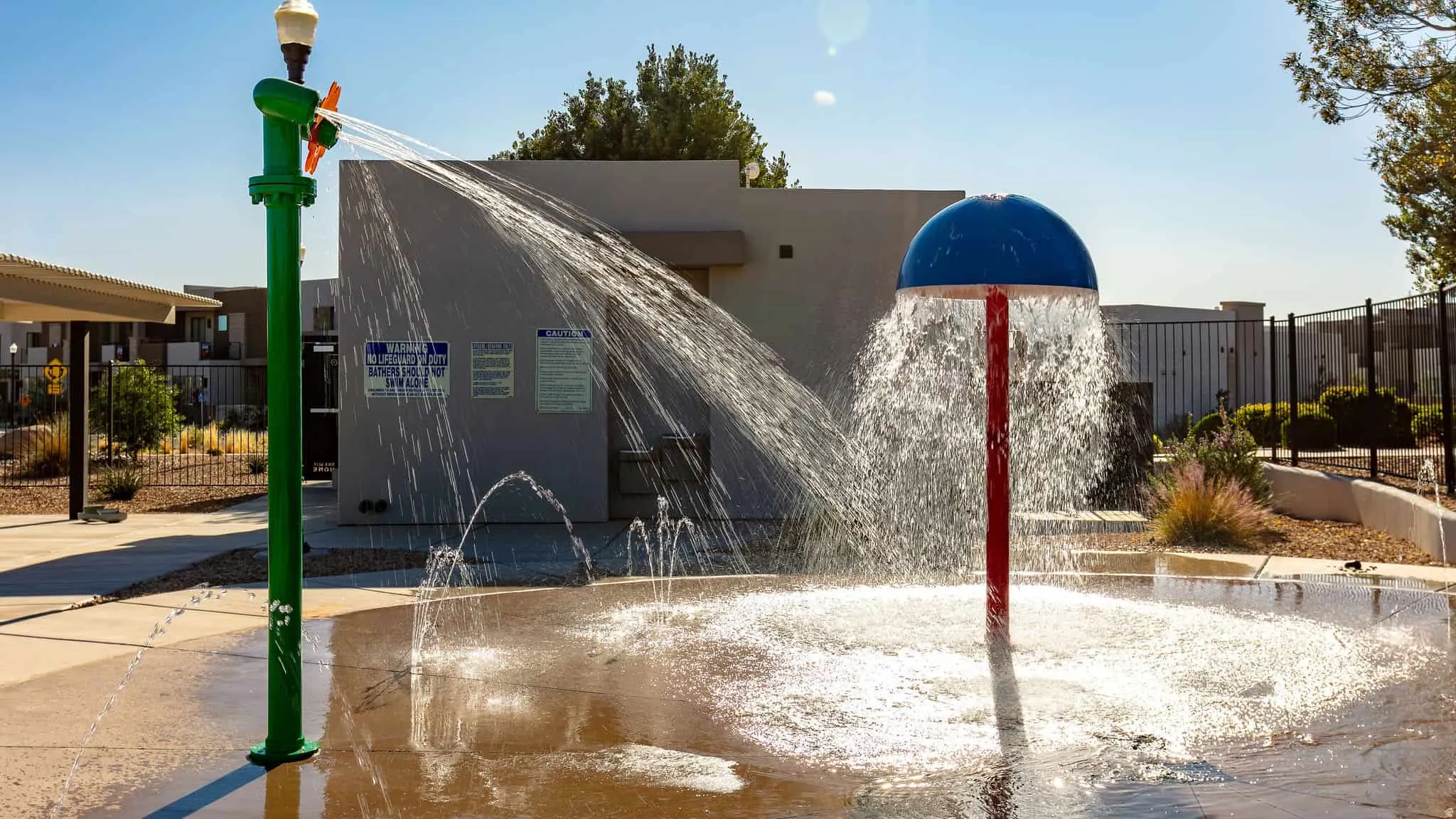 Splash pad featuring two water play structures, a green upright sprayer, and a red and blue mushroom fountain, all set on a concrete surface