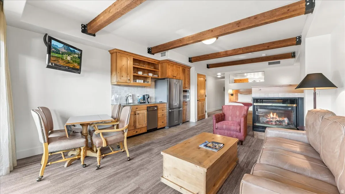 Open-concept living area with exposed wood beams, a stone-clad fireplace, and a built-in kitchen featuring stainless steel appliances and wood cabinetry