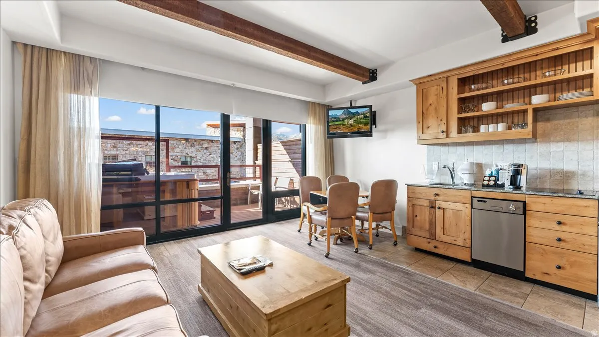 Open-concept living space featuring exposed wood beams, a full wall of glass with exterior access, wood-finish flooring, a dining area, and a kitchen with wood cabinetry and stainless steel appliances