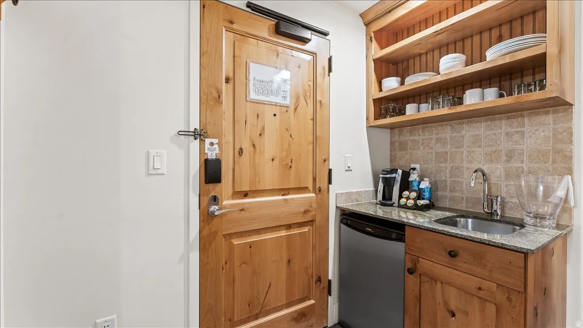 Compact wet bar with granite countertop, integrated stainless steel sink, and light wood cabinetry