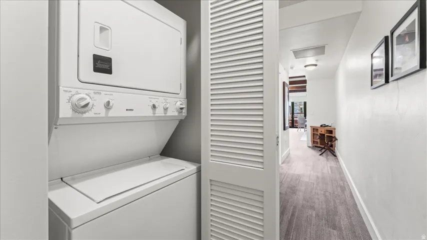 Dedicated laundry closet featuring a stacked washer and dryer, louvered bi-fold door, and wood-finish flooring extending into the hallway