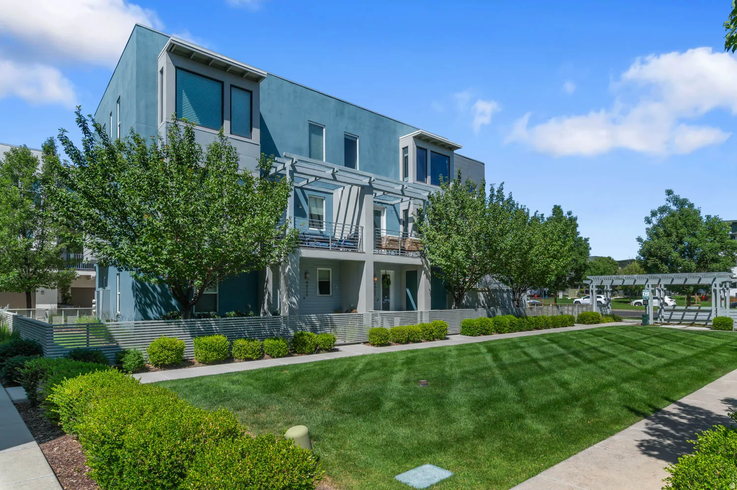 Modern multi-story building featuring a blue and gray facade, multiple balconies with metal railings, large windows, and a structured pergola