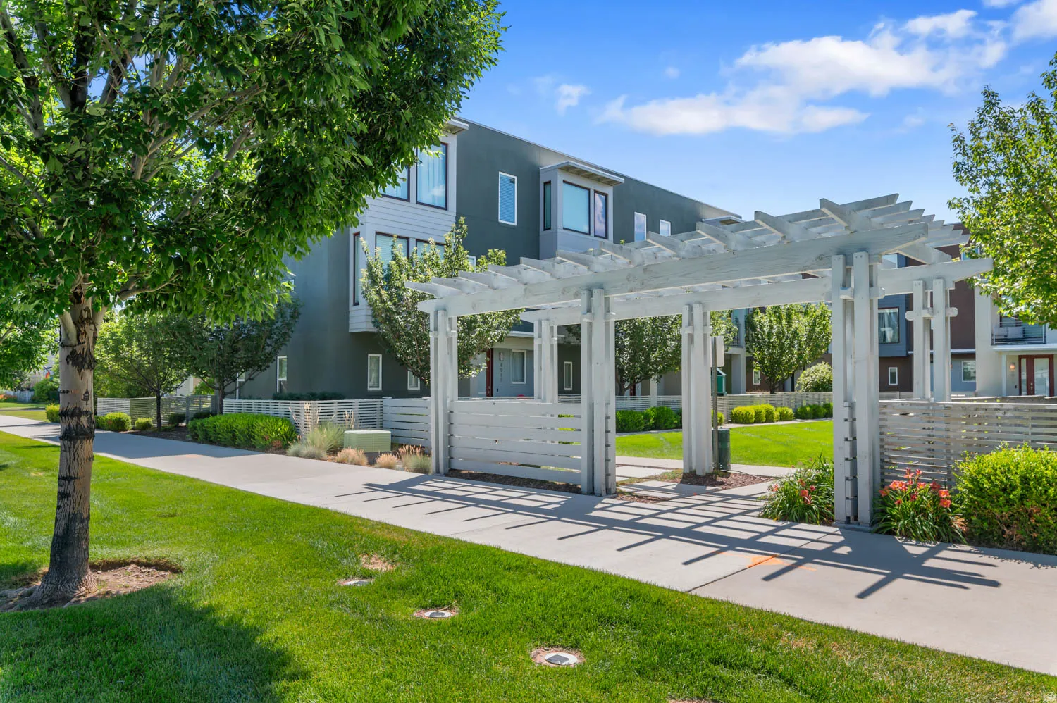 Modern multi-story building featuring dark grey siding and large windows, surrounded by a manicured lawn, mature trees, and a white pergola structure