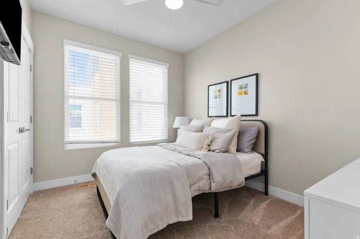 Neutral-toned room featuring a light beige carpet, two windows with blinds, a white ceiling fan, and white baseboards