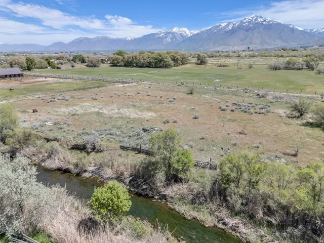 Expansive rural landscape featuring a winding river, mature trees, open fields, and distant snow-capped mountains under a bright sky