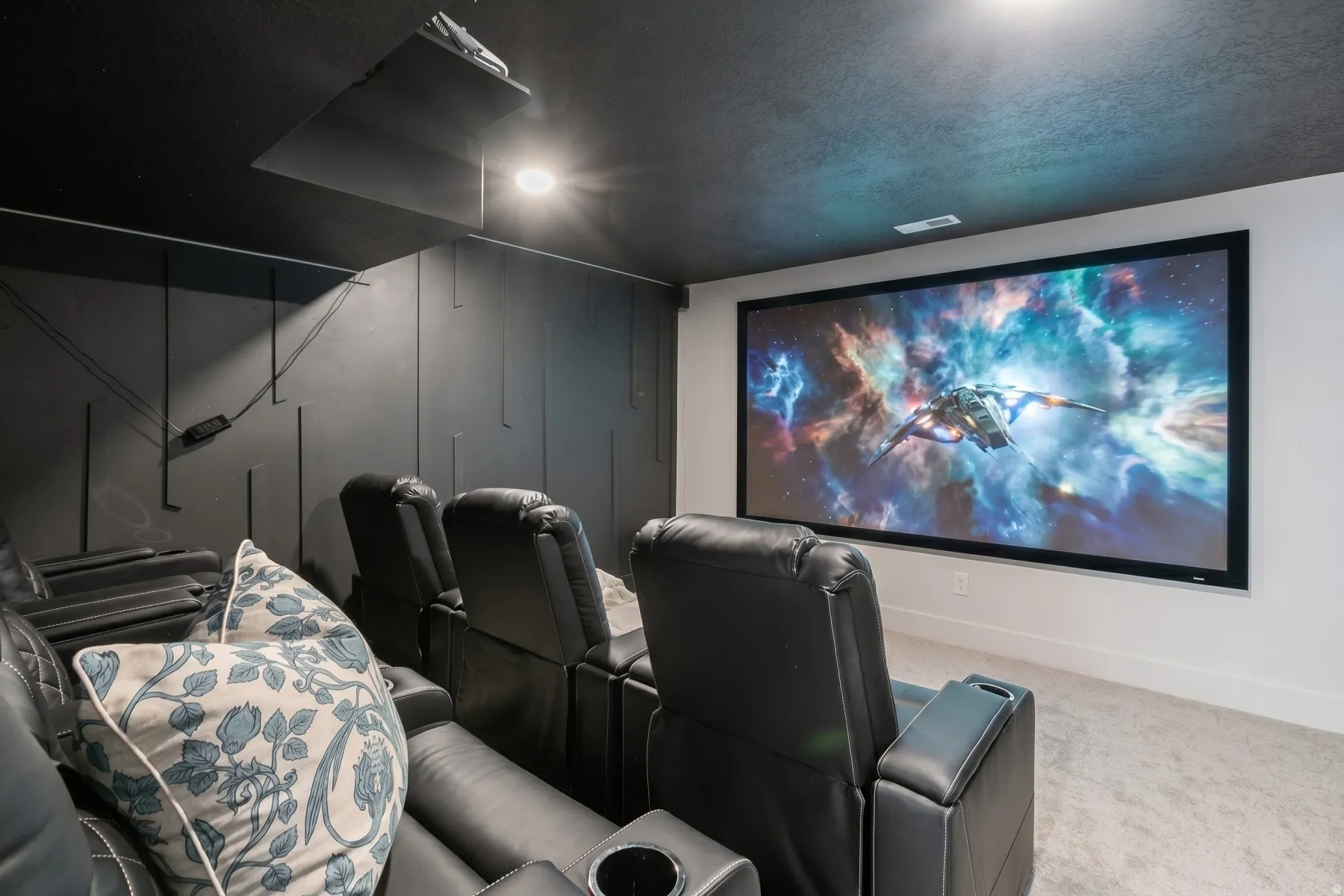 Dedicated media room featuring a large projection screen, dark textured walls, recessed ceiling lighting, and light-colored carpeting