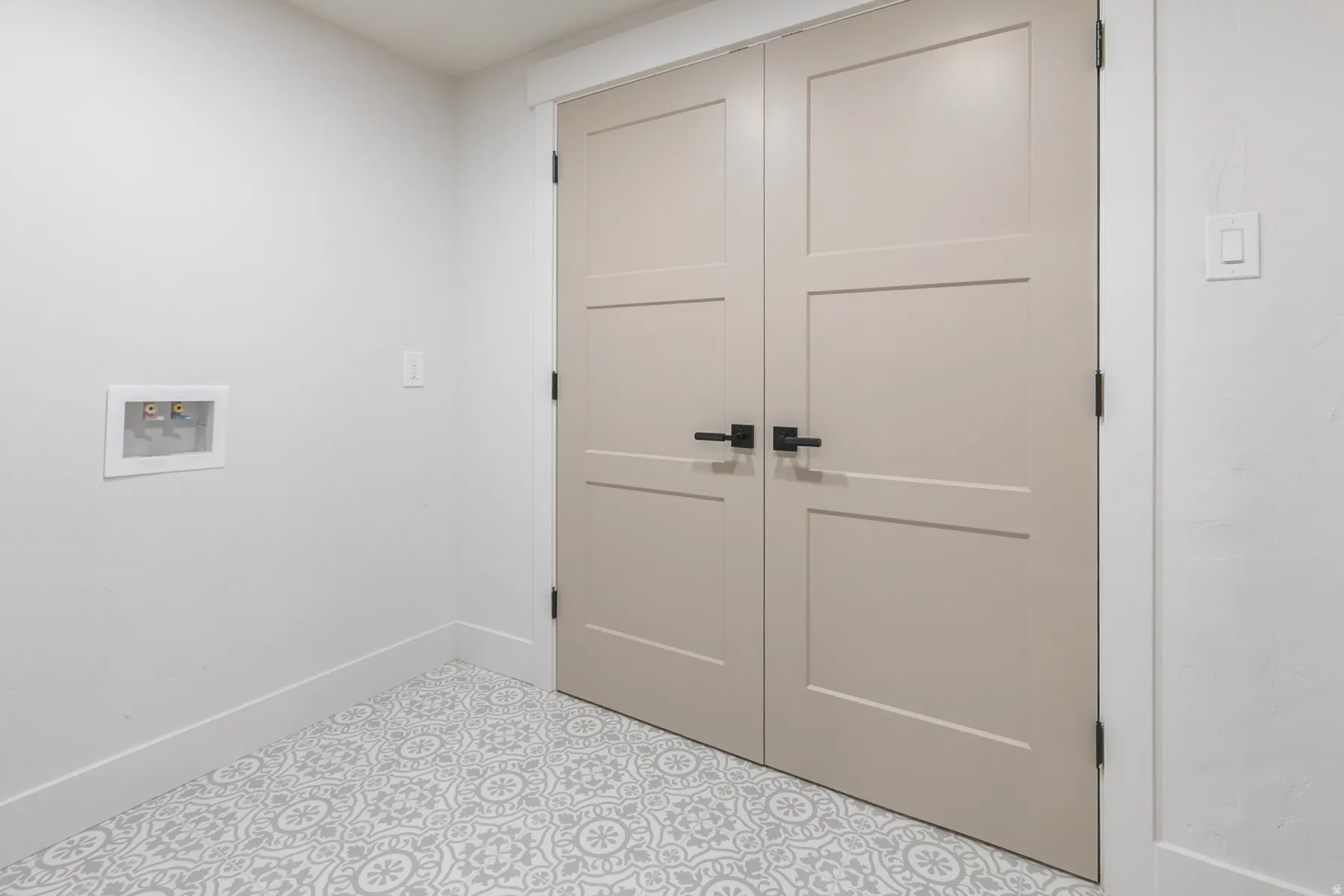 Dual panel doors with matte black hardware, patterned floor tile, white baseboards, and a recessed utility box