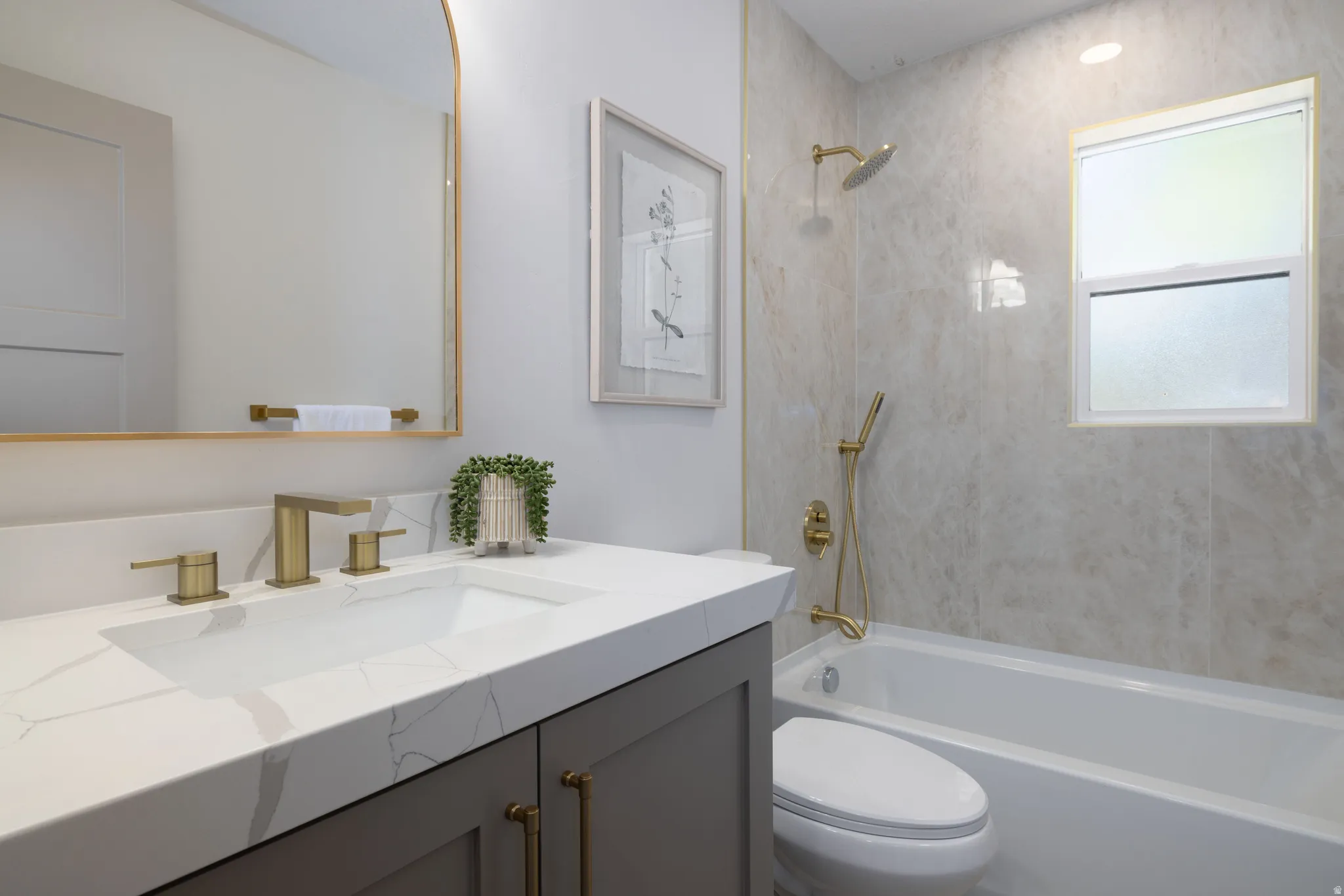 Bathroom featuring a white bathtub with gold-finish fixtures, a window, and large format wall tiling