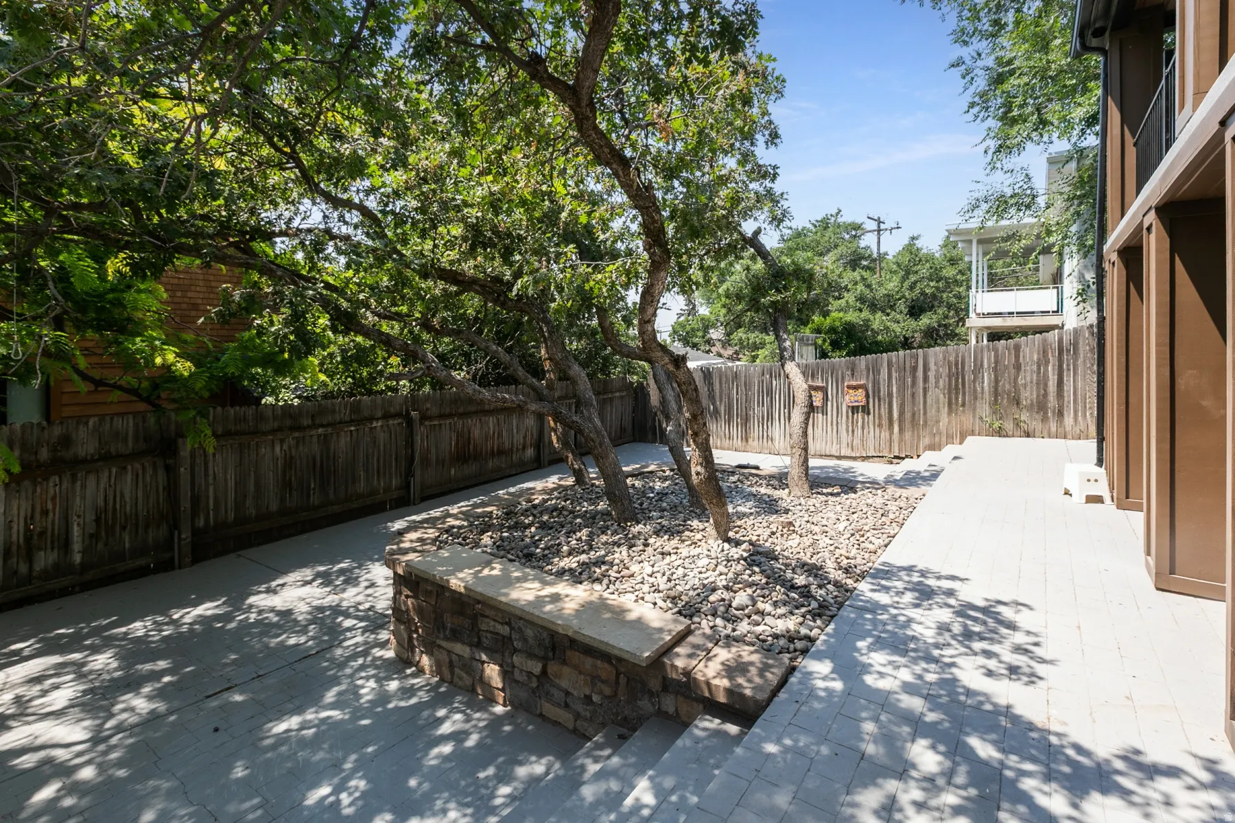 Outdoor patio area featuring light-colored pavers, mature trees, and a stone-clad raised planter with stone steps