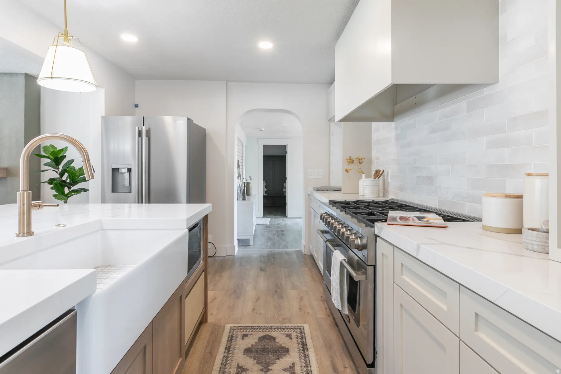 Kitchen featuring stainless steel appliances, a white farmhouse sink with a gold-tone faucet, light-toned cabinetry, light-colored countertops, and wood-finish flooring