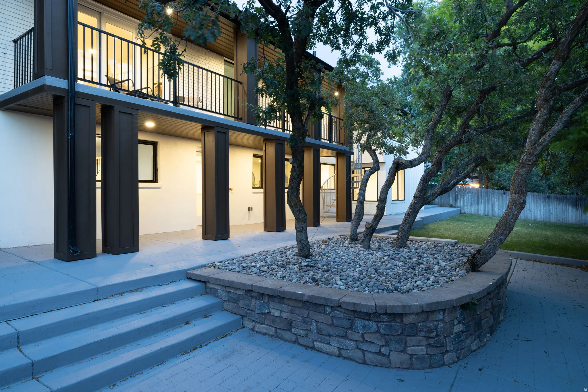 Exterior featuring a stone-faced planter with mature trees, a concrete patio, and a second-story balcony with black railings