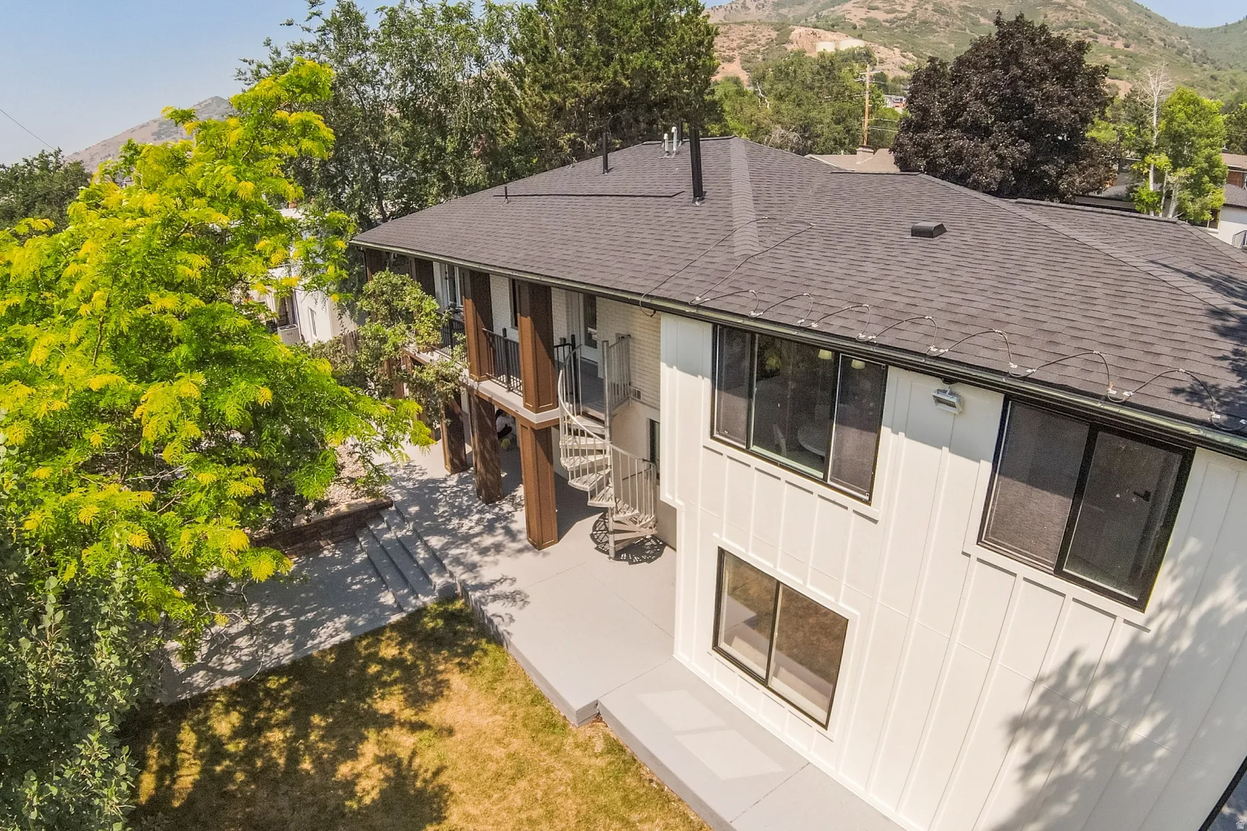 Contemporary exterior featuring white siding, a dark shingle roof, dark-framed windows, a spiral staircase, and a mountain backdrop