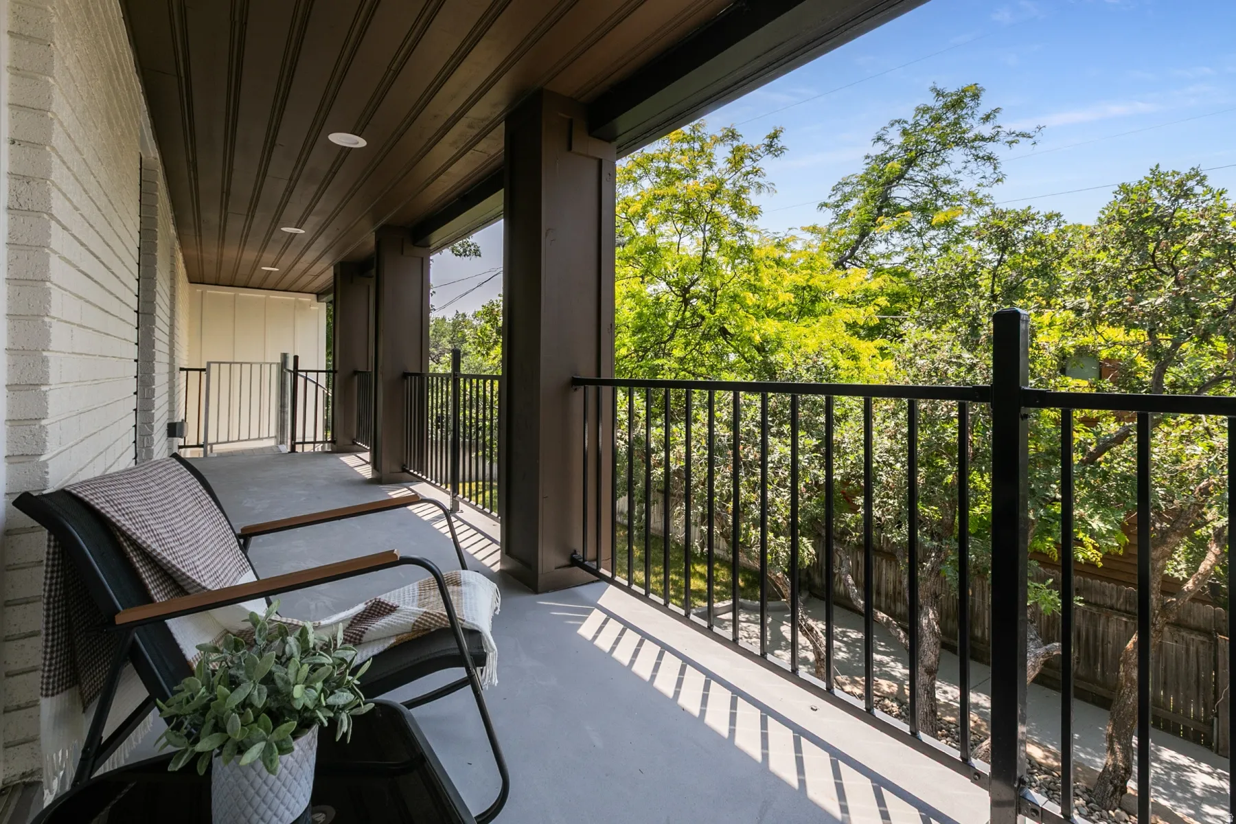 Expansive covered balcony featuring a textured white brick wall, dark wood-finish ceiling, recessed lighting, and black metal railings, overlooking mature trees