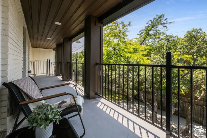 Expansive covered balcony featuring a textured white brick wall, dark wood-finish ceiling, recessed lighting, and black metal railings, overlooking mature trees