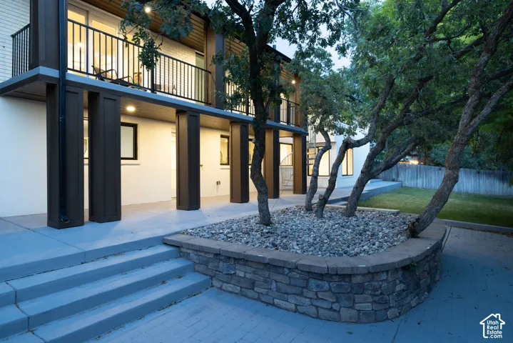Exterior featuring a stone-faced planter with mature trees, a concrete patio, and a second-story balcony with black railings