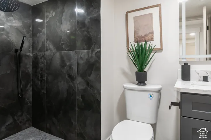 Bathroom featuring dark-toned tiled shower, rain shower head, hand-held shower, white toilet, and vanity with dark cabinetry