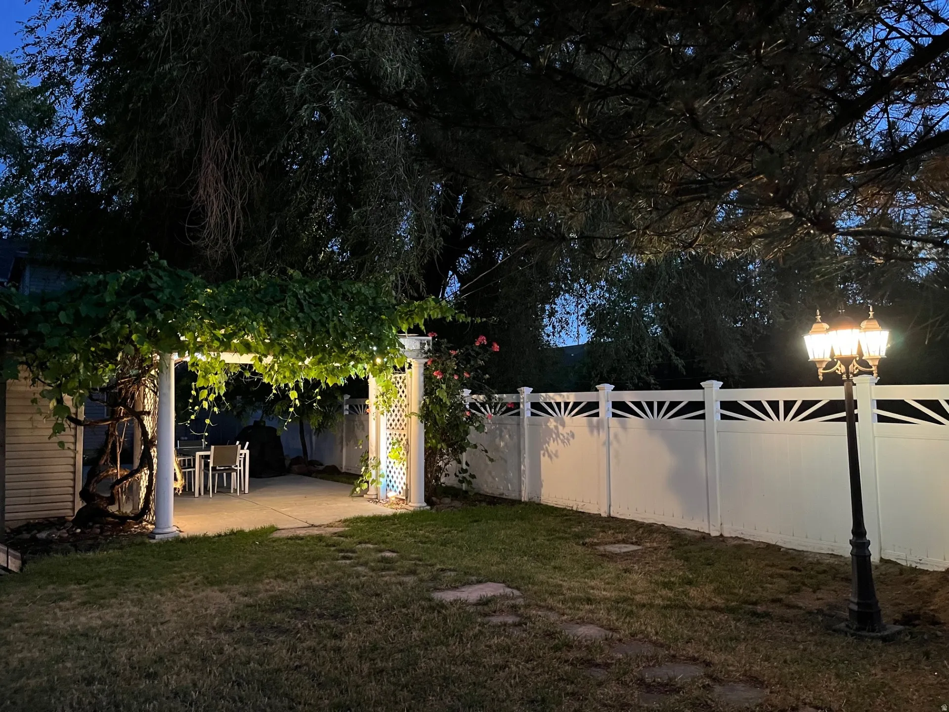 Backyard featuring a covered patio with string lights, a white privacy fence with decorative toppers, and a classic black lamppost