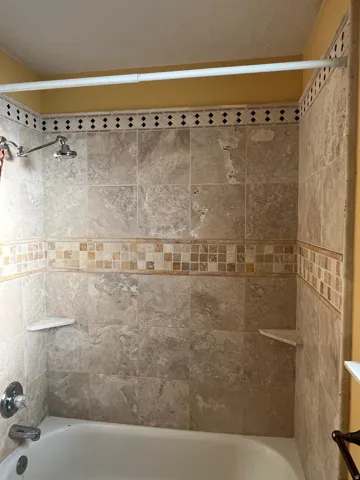 Bathtub with tiled surround featuring mosaic accents, an adjustable shower head, and built-in corner shelving
