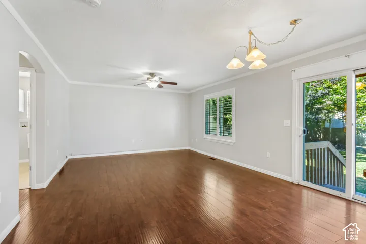 Spacious room featuring wood-finish flooring, white baseboards and crown molding, a ceiling fan with light fixture, plantation shutters on the window, and a sliding glass door leading to an outdoor deck and green space