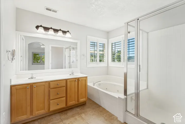 Bathroom featuring a dual vanity with light wood cabinetry, a large framed mirror, a separate shower enclosure, a built-in jetted tub, and multiple windows with plantation shutters