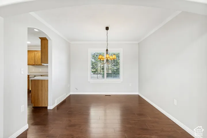 Spacious room featuring wood-finish flooring, a contemporary chandelier, a window with horizontal blinds, and an arched doorway opening to a kitchen with wood cabinetry