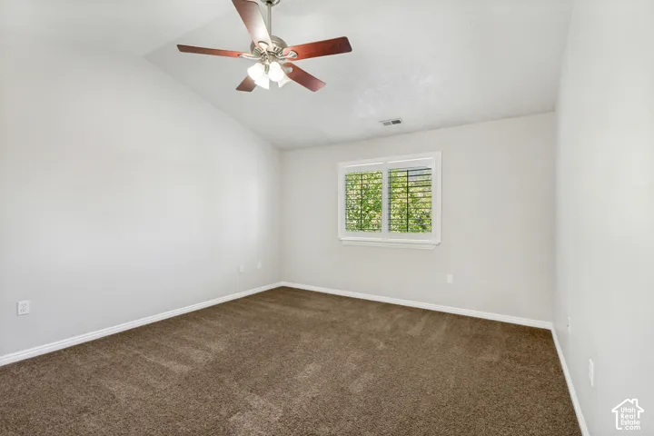 Spacious primary room featuring vaulted ceilings, a ceiling fan with integrated lighting, a window with plantation shutters, and brown carpeting