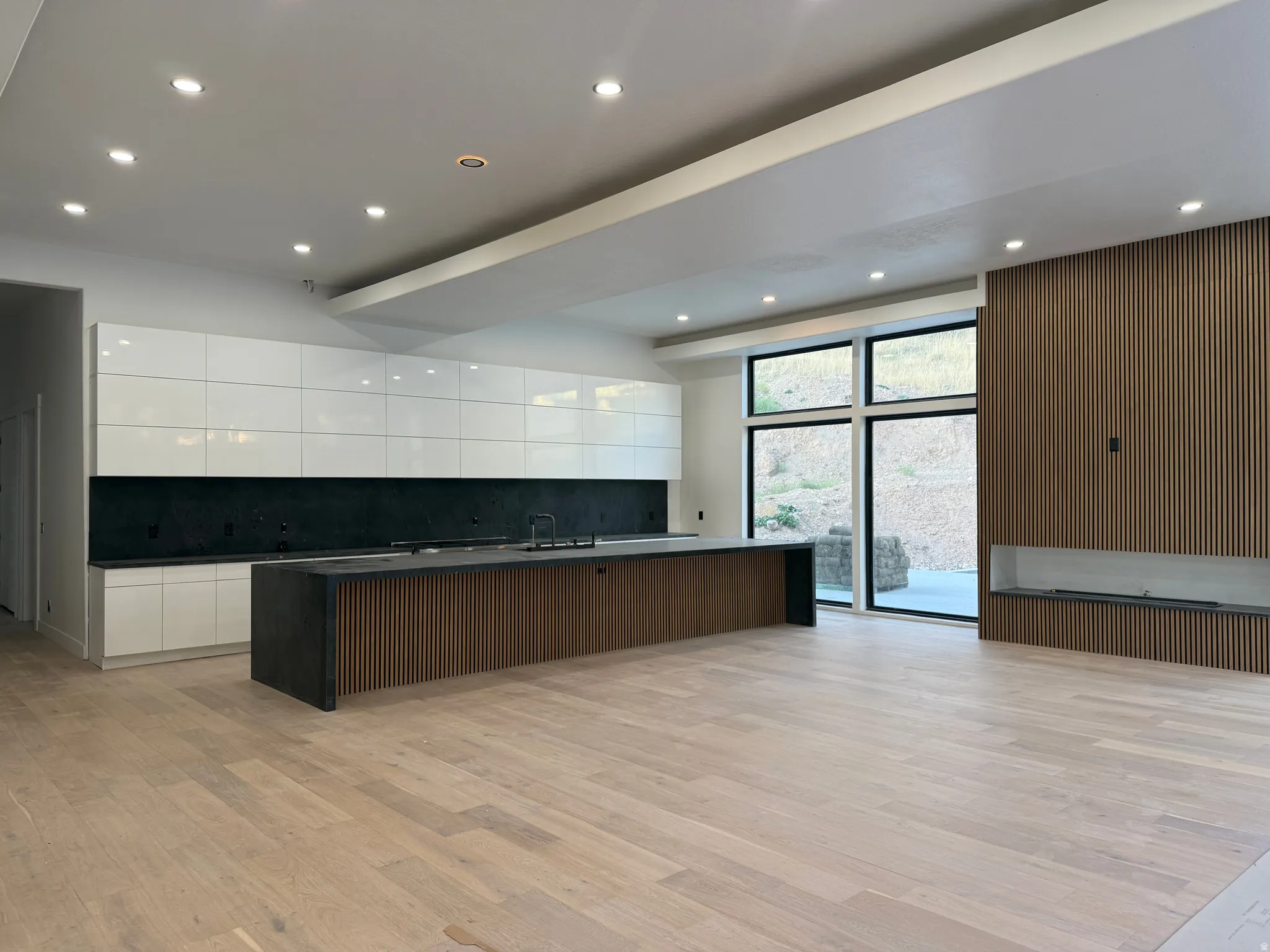 Open-concept living area with light wood-finish flooring, a contemporary kitchen island featuring a dark countertop and slatted wood-finish base, white upper and lower cabinetry with a dark backsplash, a built-in fireplace, and large windows providing abu